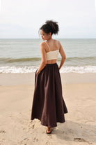 Woman standing on a beach wearing a beige top and brown skirt.