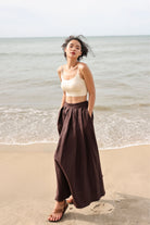 Woman standing on a beach wearing a beige top and brown skirt.
