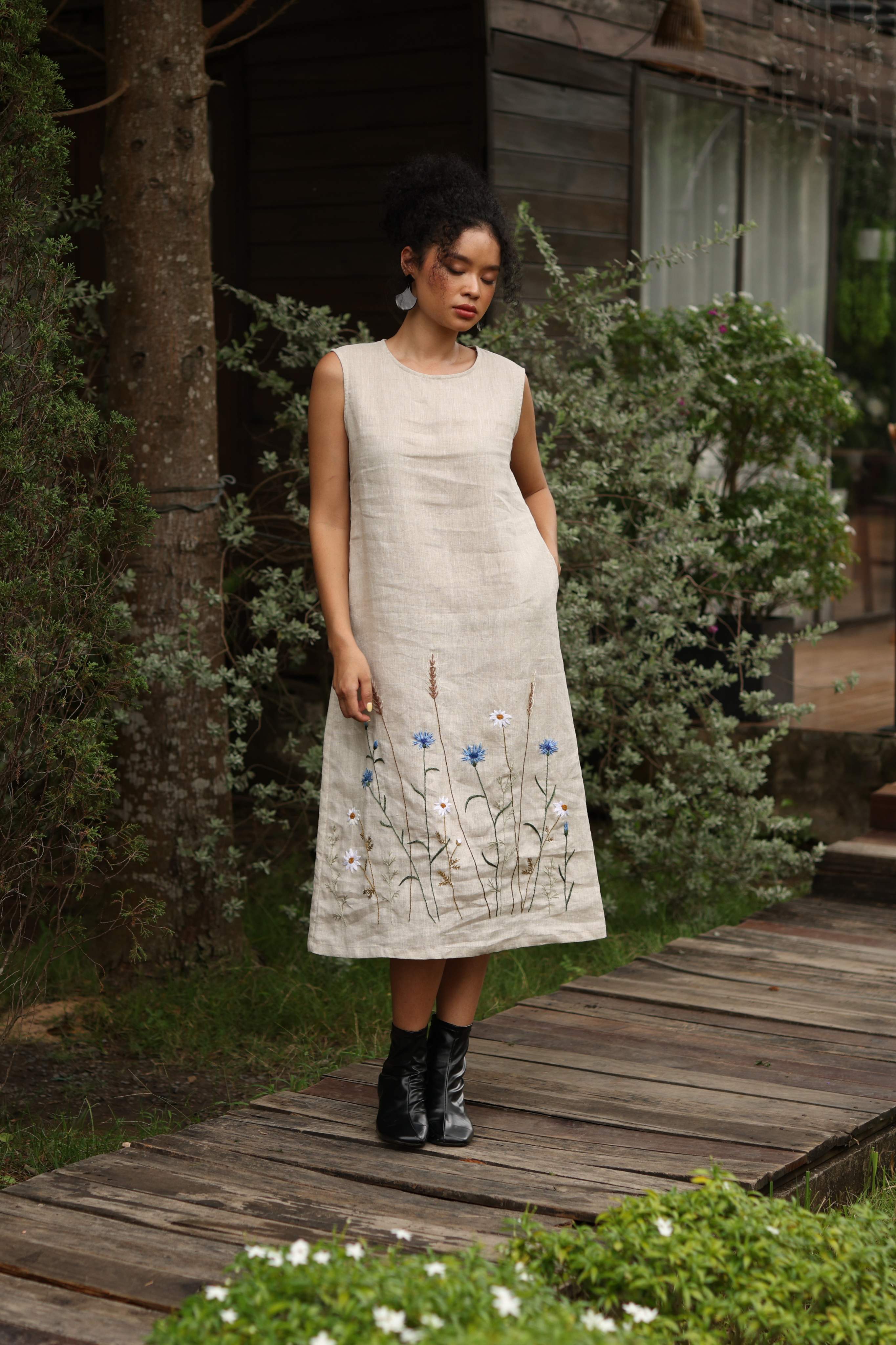 Woman in a light-colored dress with floral embroidery standing on a wooden path in a garden.