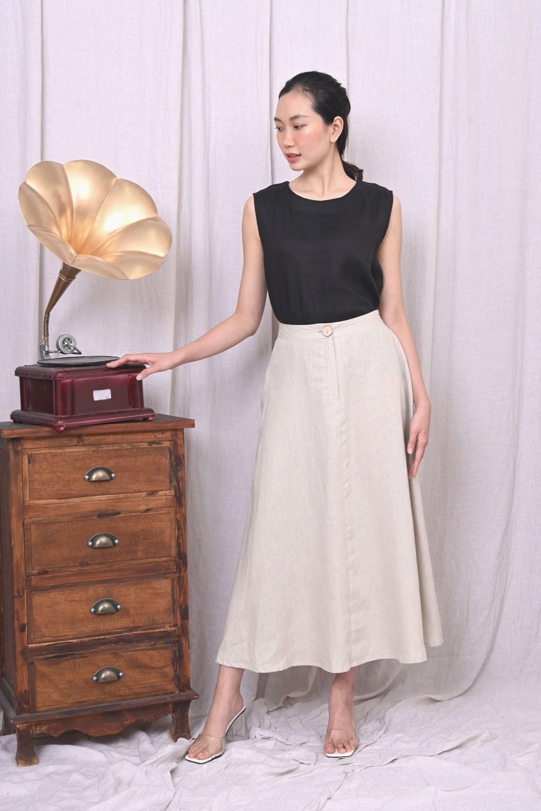Woman in black top and white skirt standing next to a wooden gramophone cabinet against a white curtain.