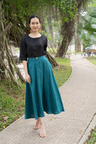 Woman in black top and teal skirt standing on a pathway with greenery