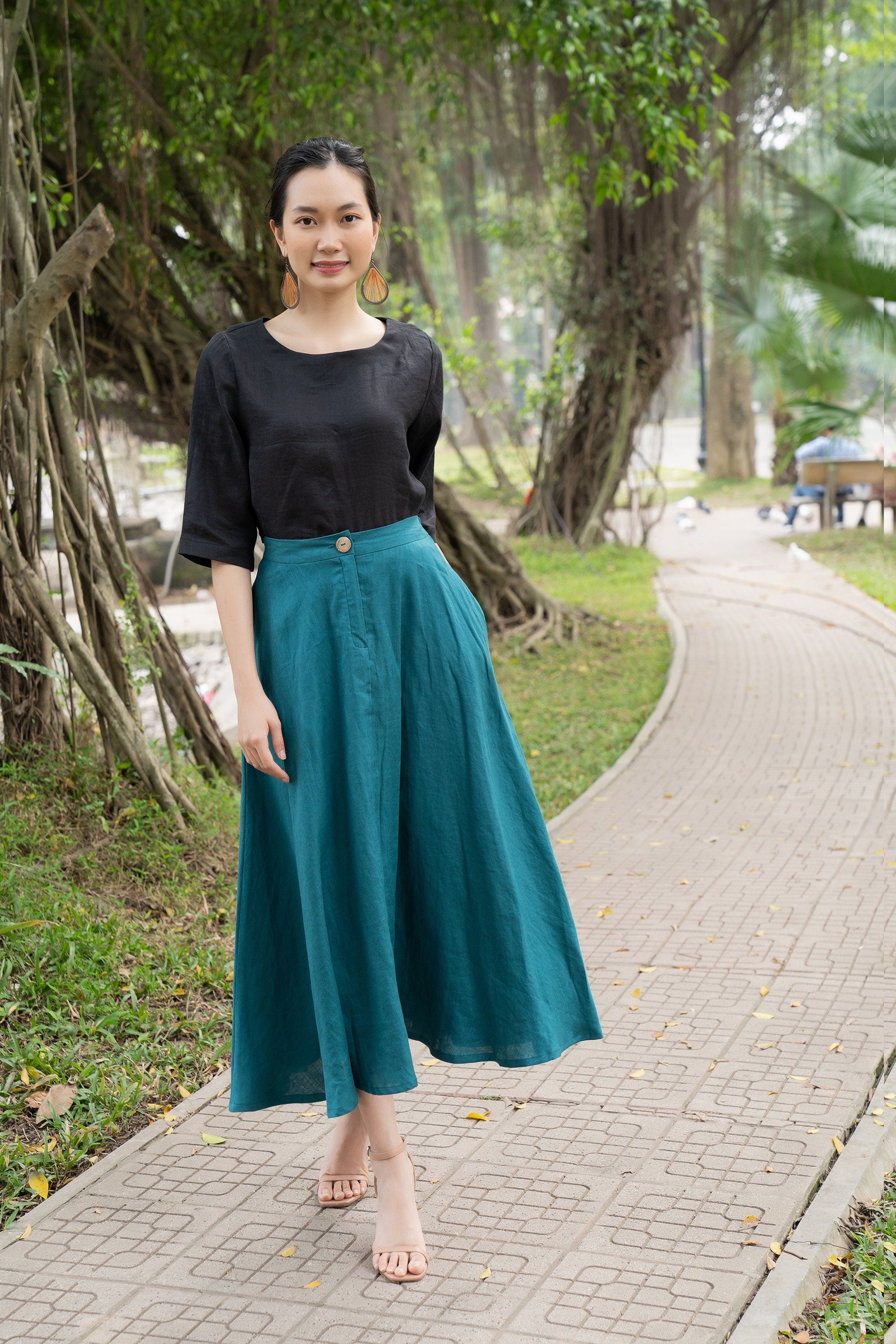 Woman in black top and teal skirt standing on a pathway with greenery