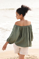 Woman wearing a green off-shoulder top and white shorts on a beach.
