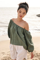 Woman wearing an off-shoulder green top and white pants on a beach.
