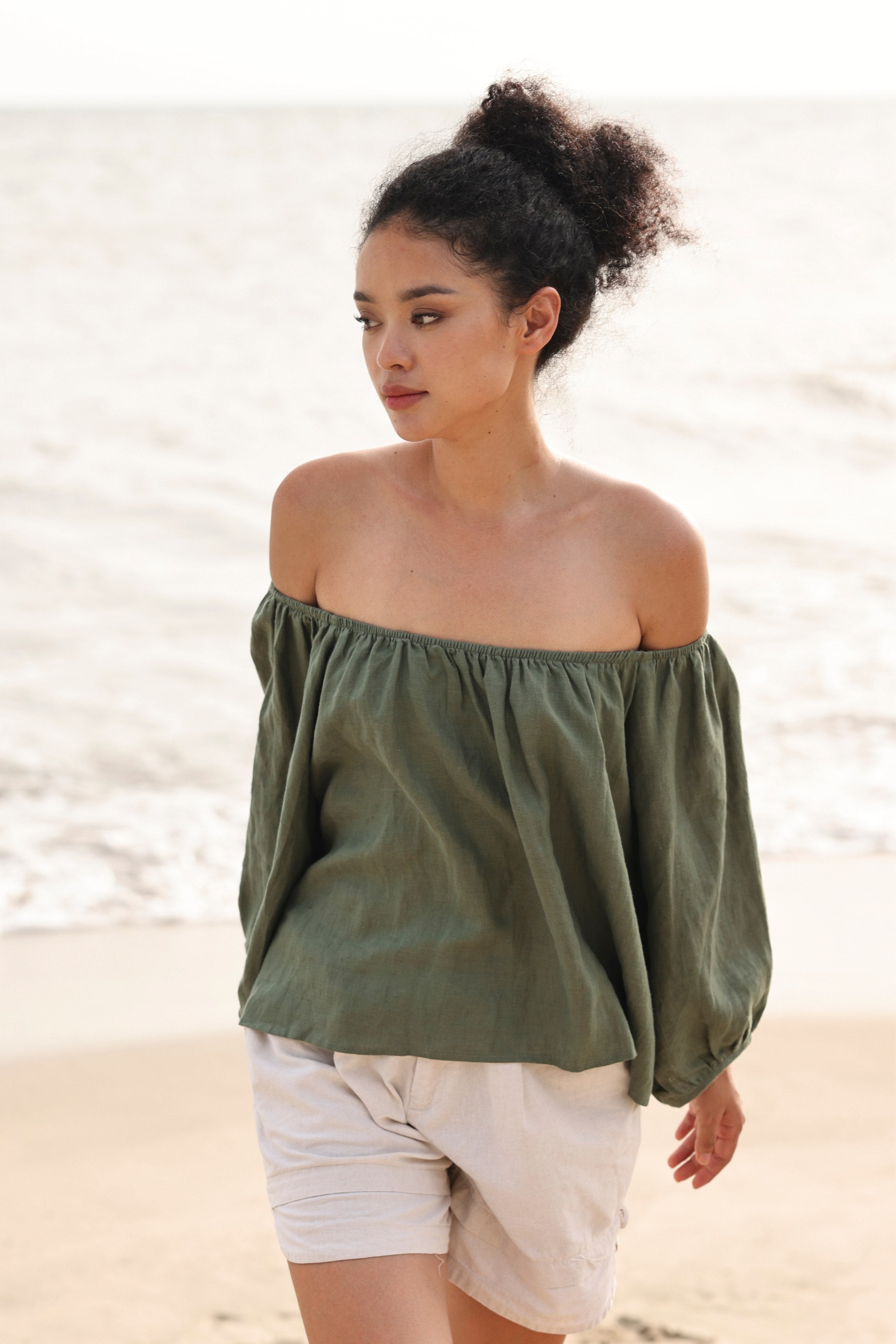 Woman wearing an off-shoulder green top and white shorts on a beach.