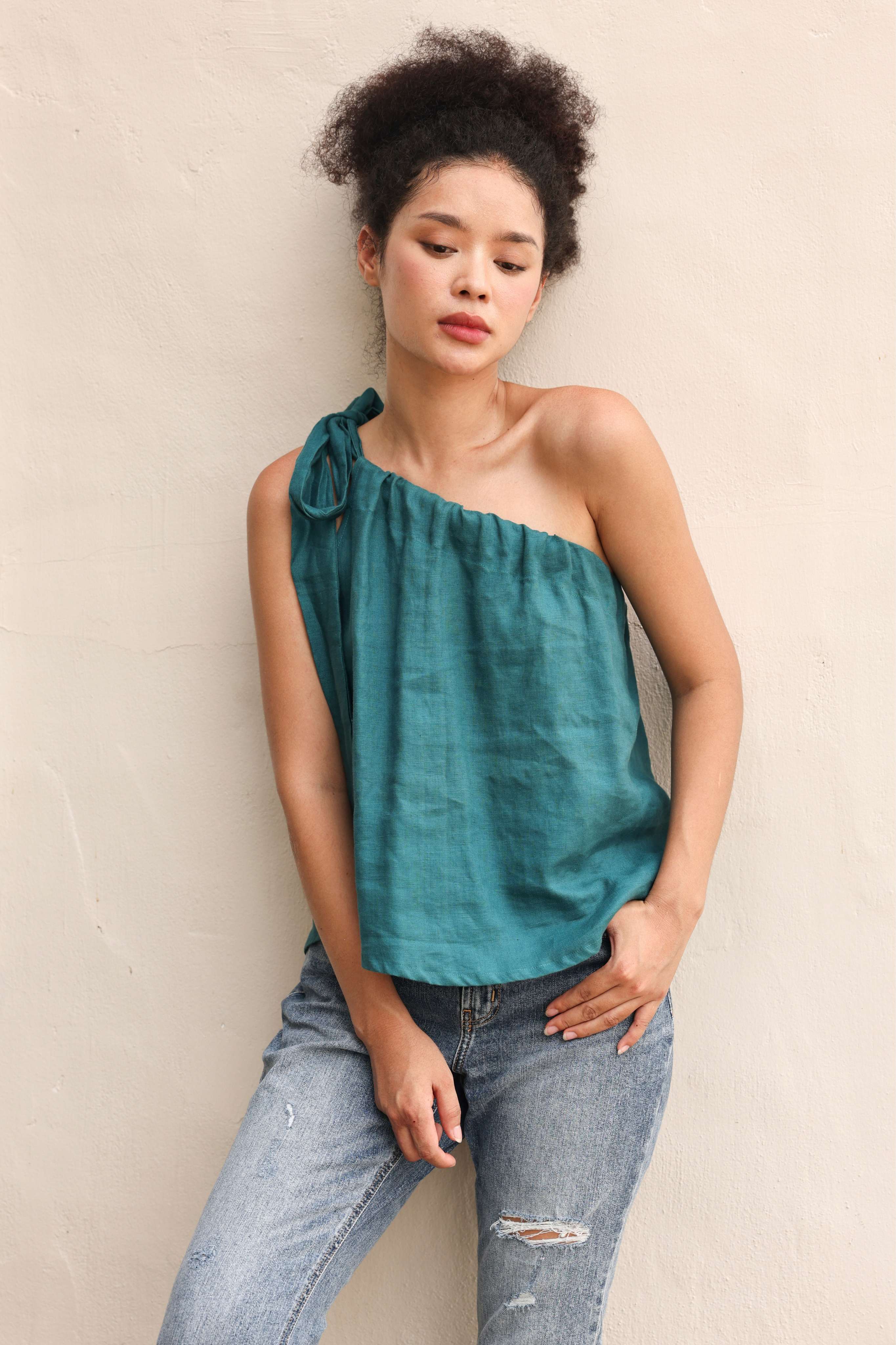 Woman wearing a teal one-shoulder top and jeans against a beige background