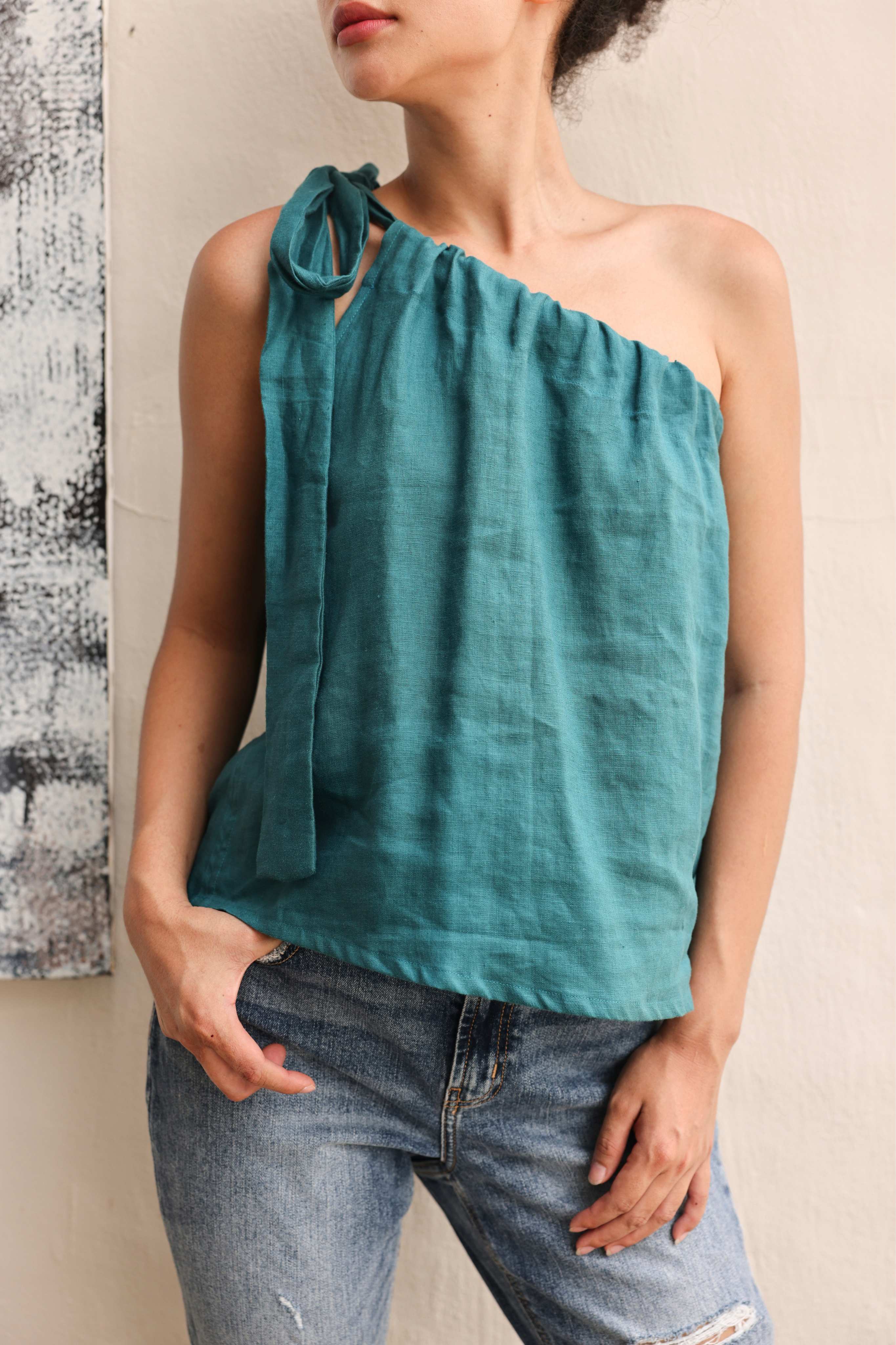 Woman wearing a teal one-shoulder top and jeans against a neutral background