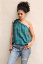 Woman wearing a teal one-shoulder top and blue jeans against a beige wall.