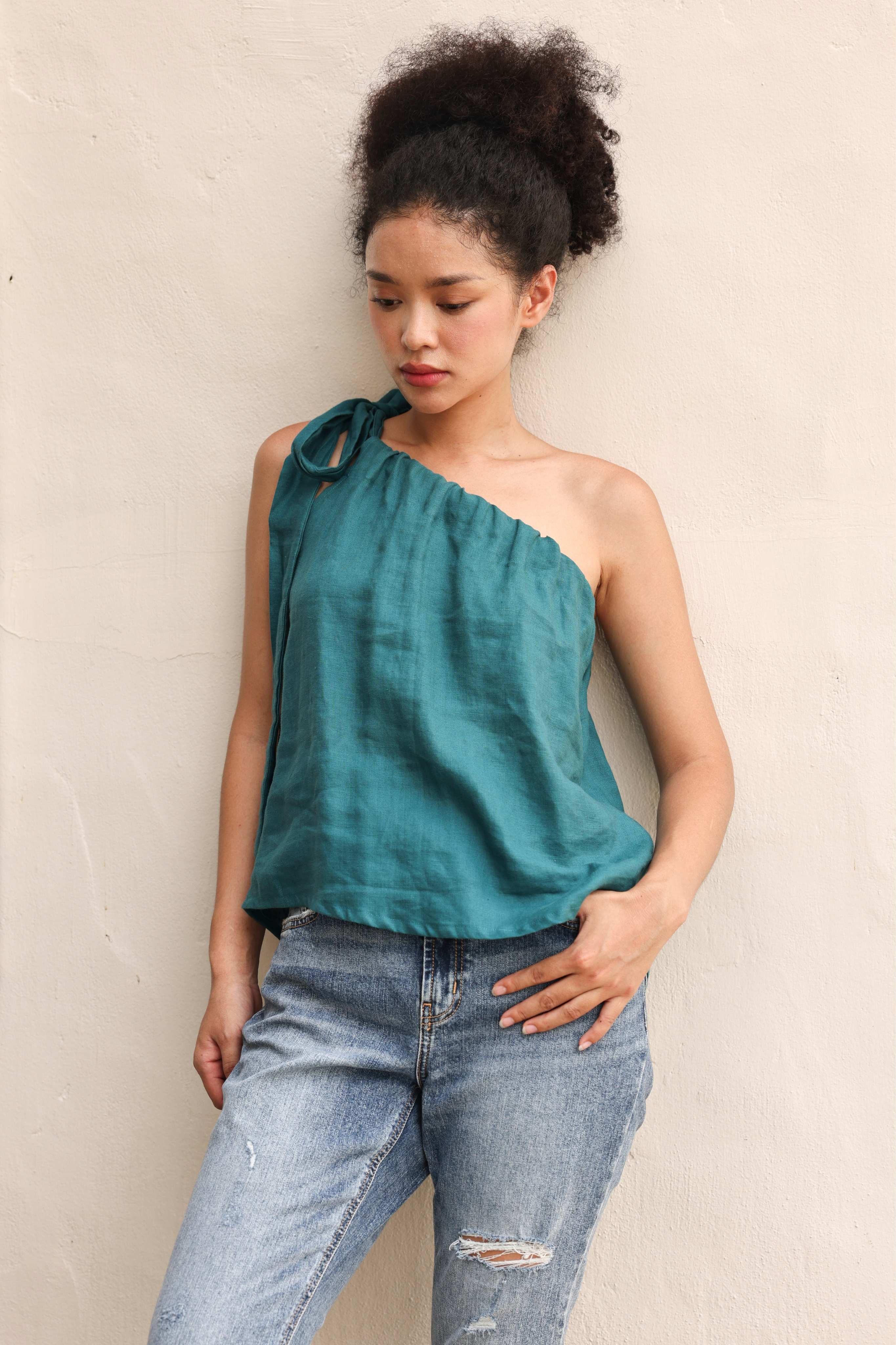 Woman wearing a teal one-shoulder top and blue jeans against a beige wall.