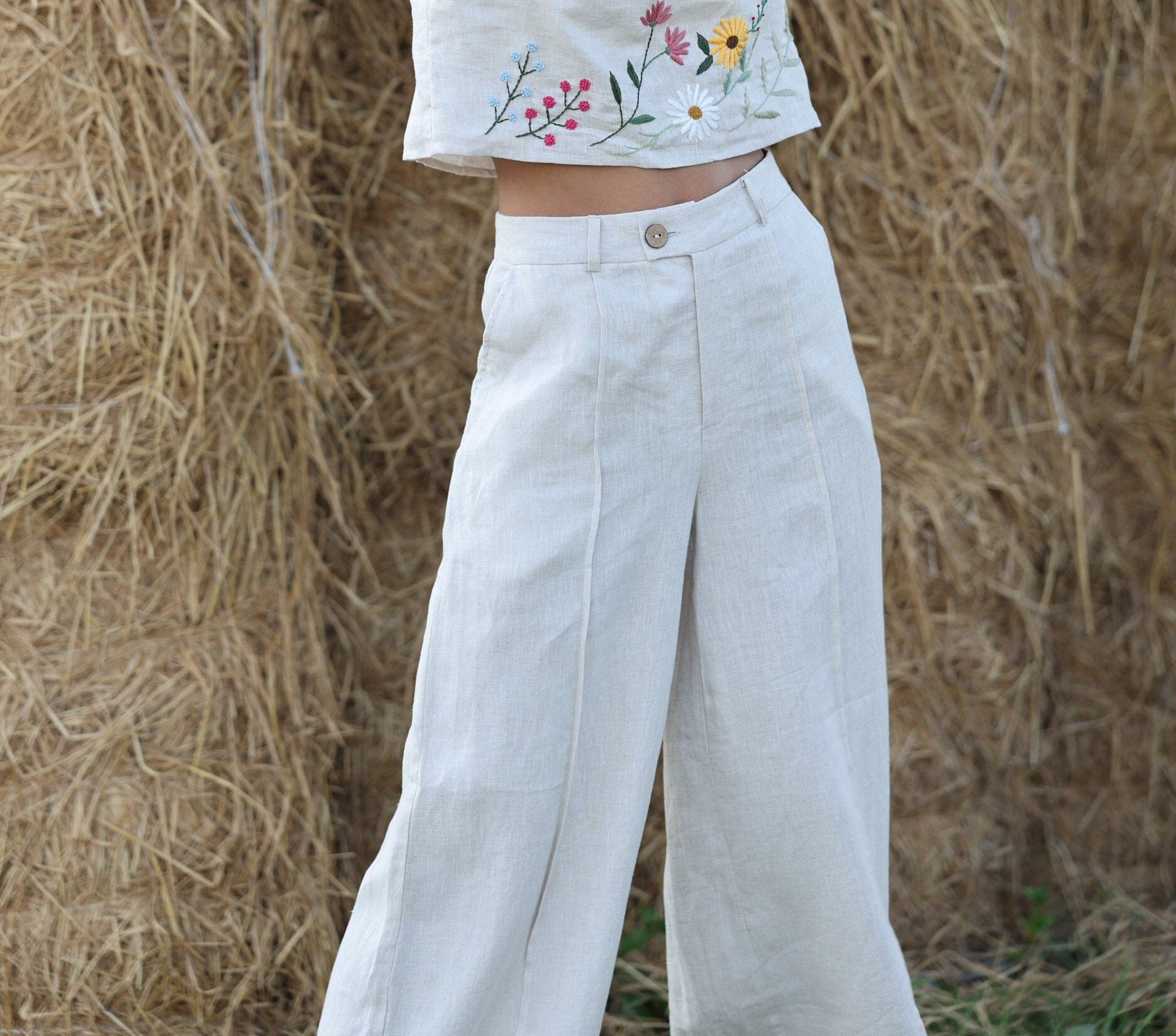 Person wearing a white embroidered top and light-colored pants standing in front of hay bales.