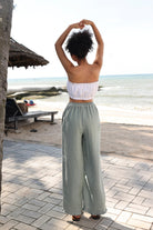 Woman in a white top and light green pants standing on a beach path with ocean view.