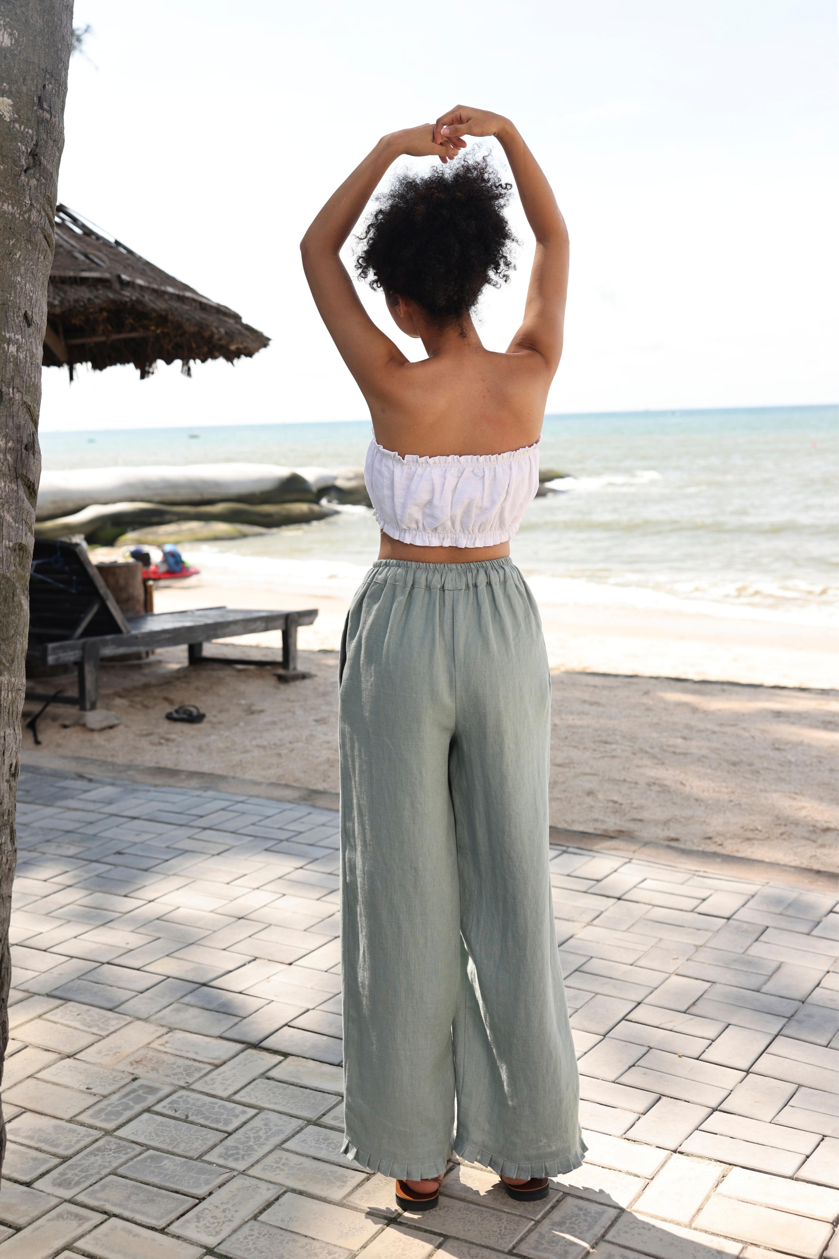 Woman in a white top and light green pants standing on a beach path with ocean view.