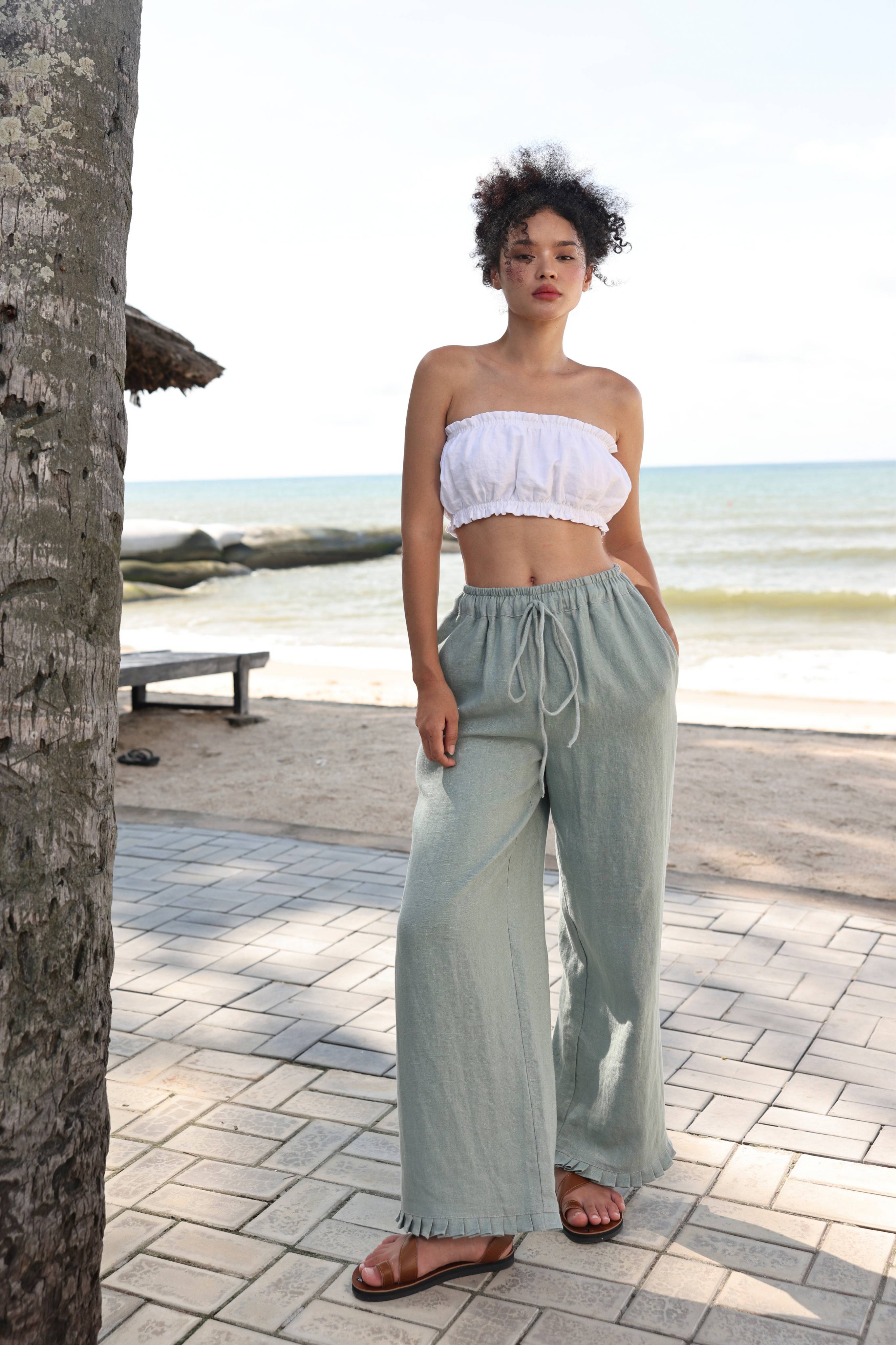 Woman in white top and pants standing on a beach path with ocean view.