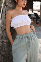 Woman wearing a white strapless top and light green pants leaning against a tree.