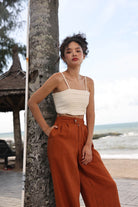 Woman in a beige top and rust-colored pants leaning against a tree by the beach.