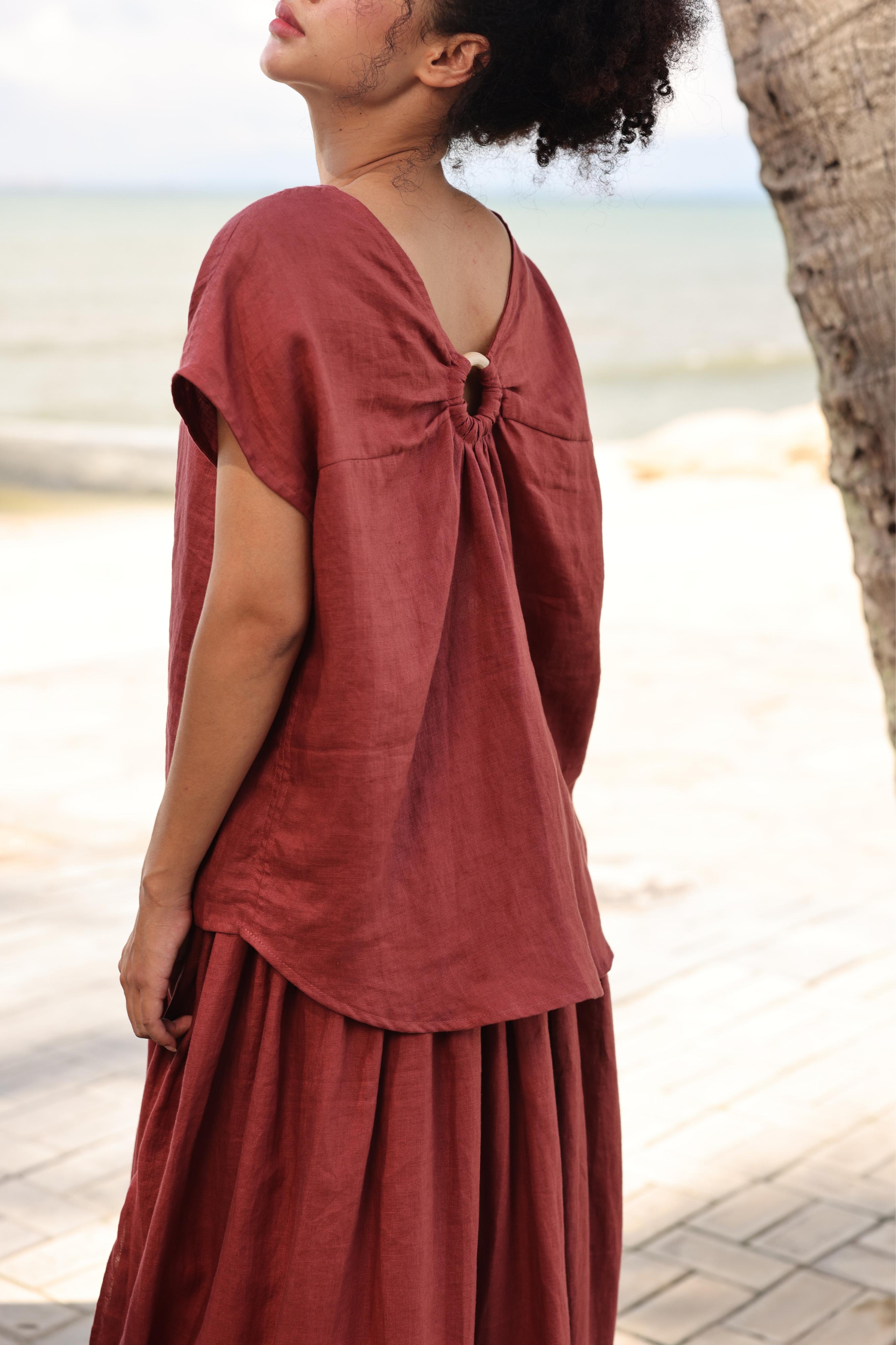 Woman wearing a rust-colored top standing by a tree on a beach.