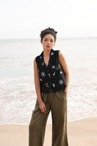 Woman wearing a black embroidered vest and green pants standing on a beach.