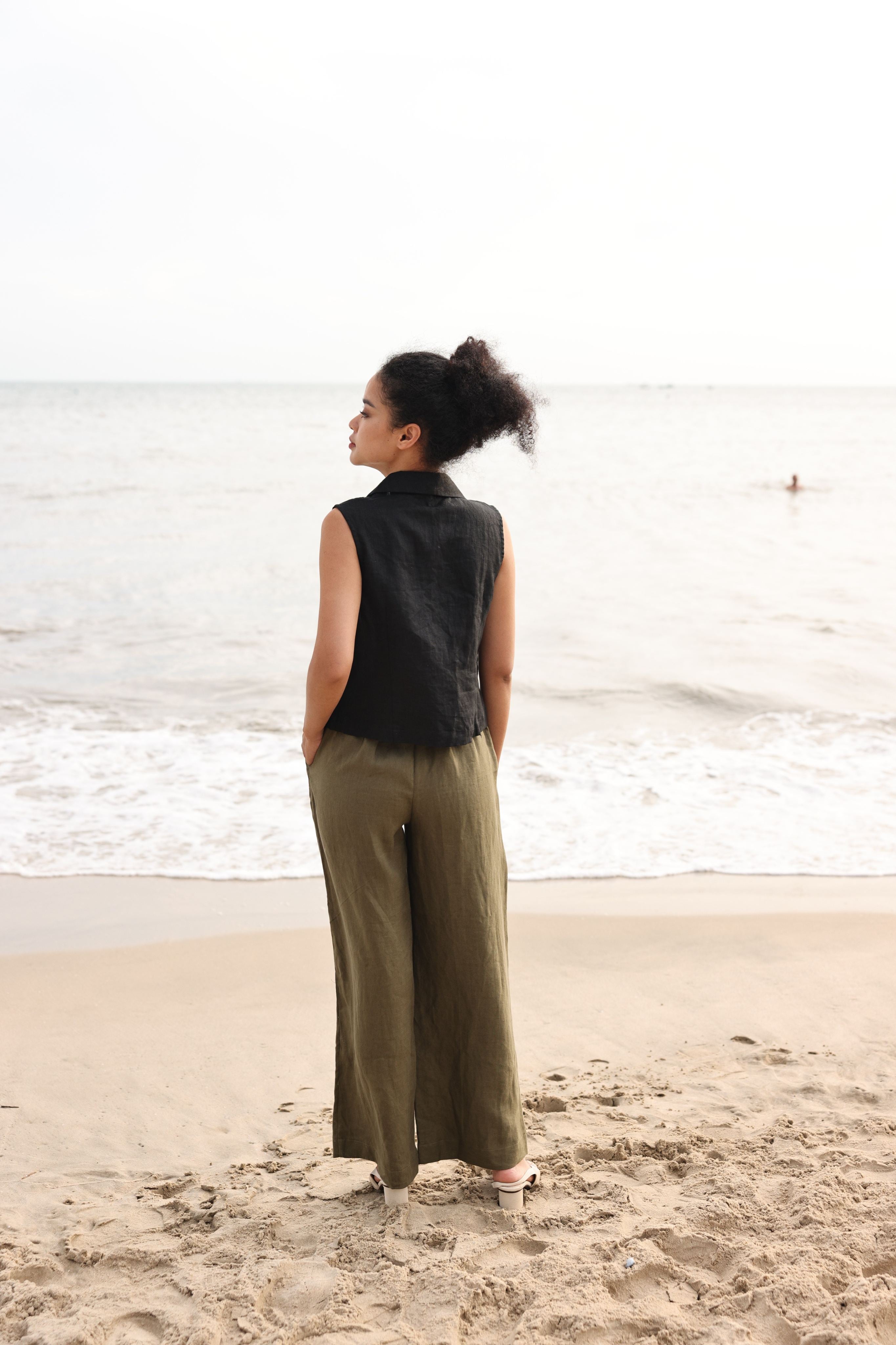 Woman standing on a beach wearing a black sleeveless top and green pants.