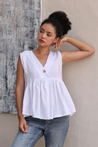 Woman wearing a white sleeveless top with a button and blue jeans against a textured wall.