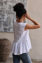 Person wearing a white sleeveless top with ruffled hem and blue jeans, standing in a room with a textured wall and large vase.