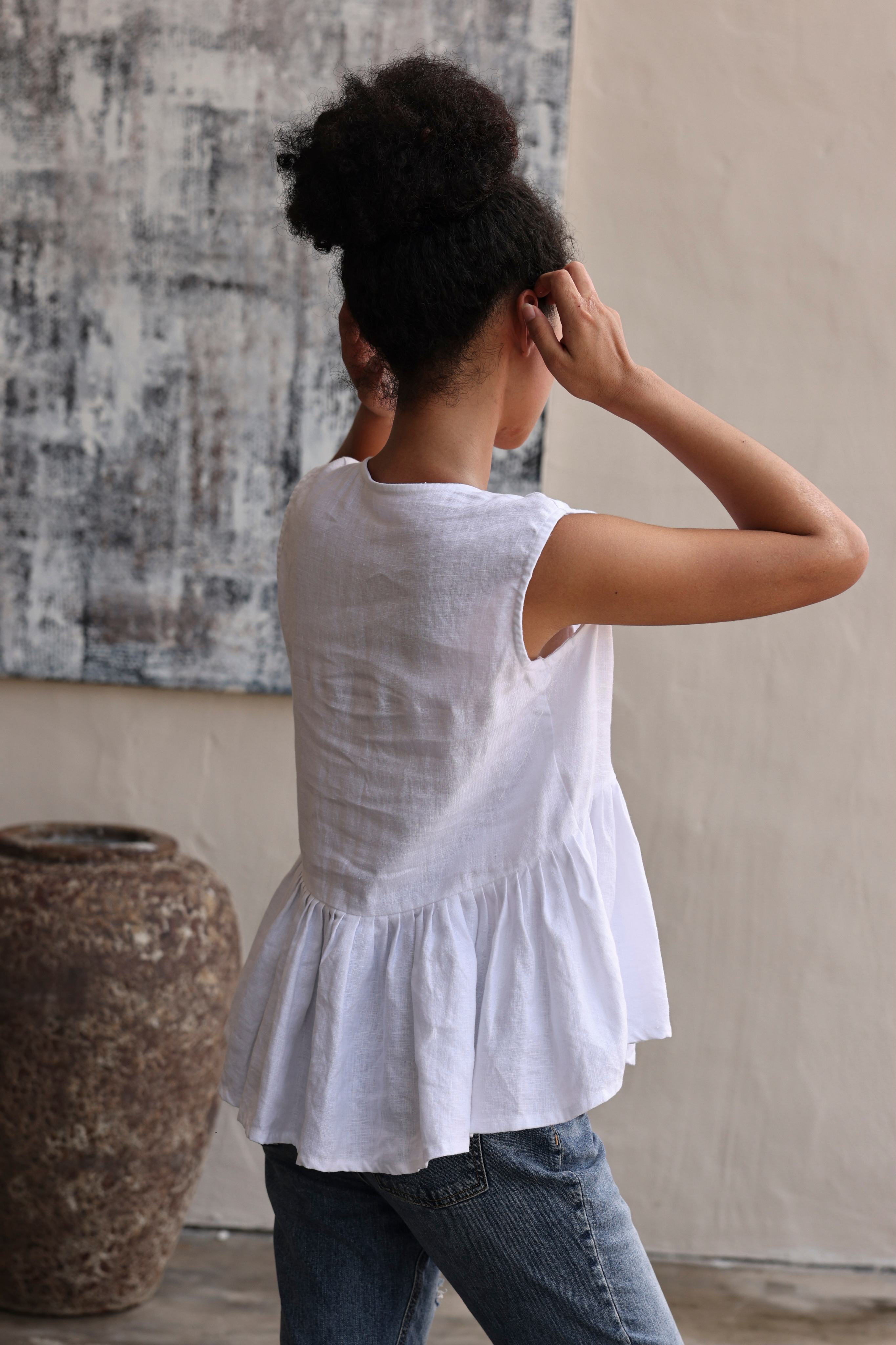Person wearing a white sleeveless top with ruffled hem and blue jeans, standing in a room with a textured wall and large vase.