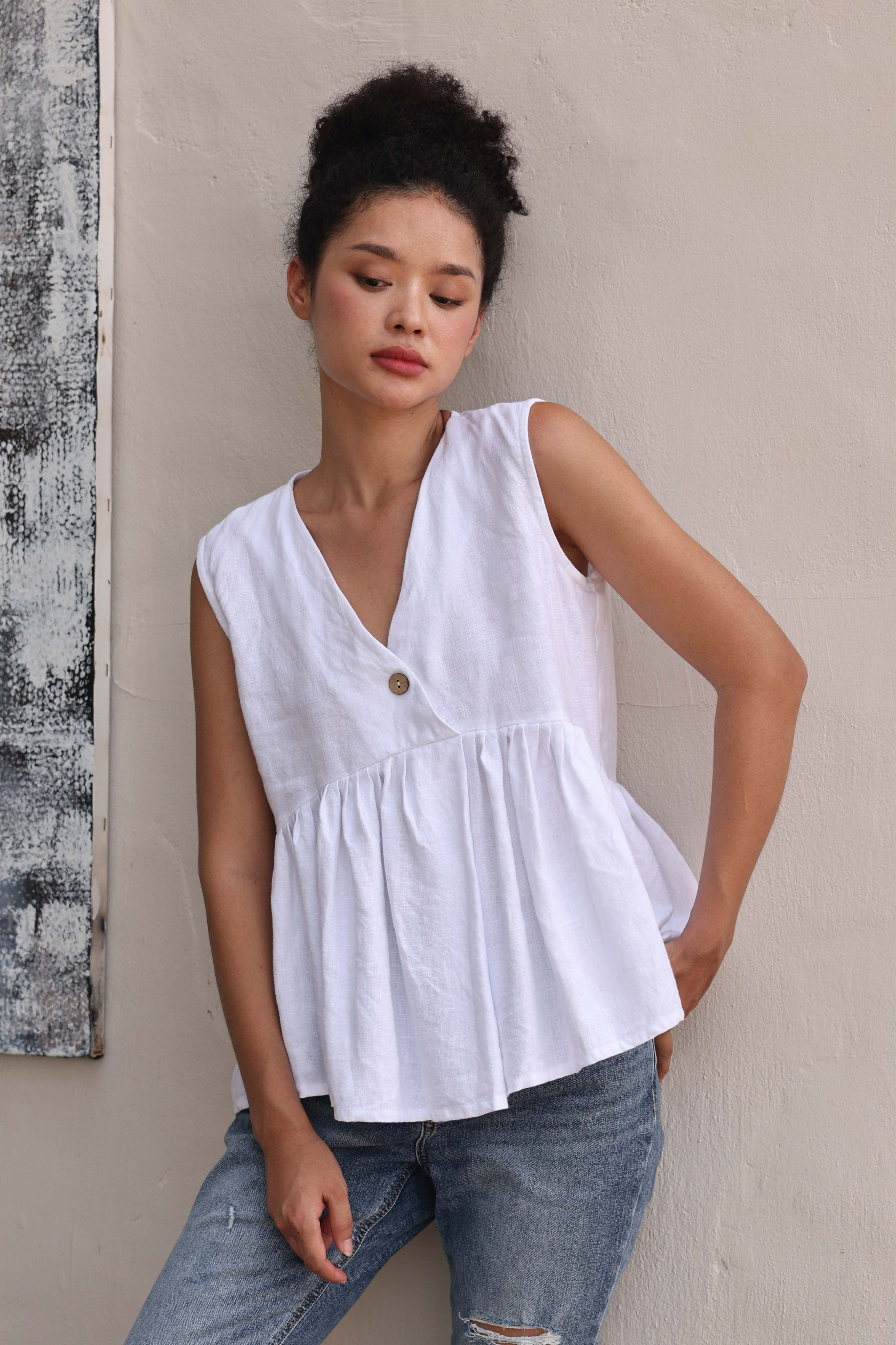 Woman wearing a white sleeveless top and blue jeans against a neutral wall.