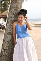 Woman in a blue top and white skirt standing by a tree on a beach.