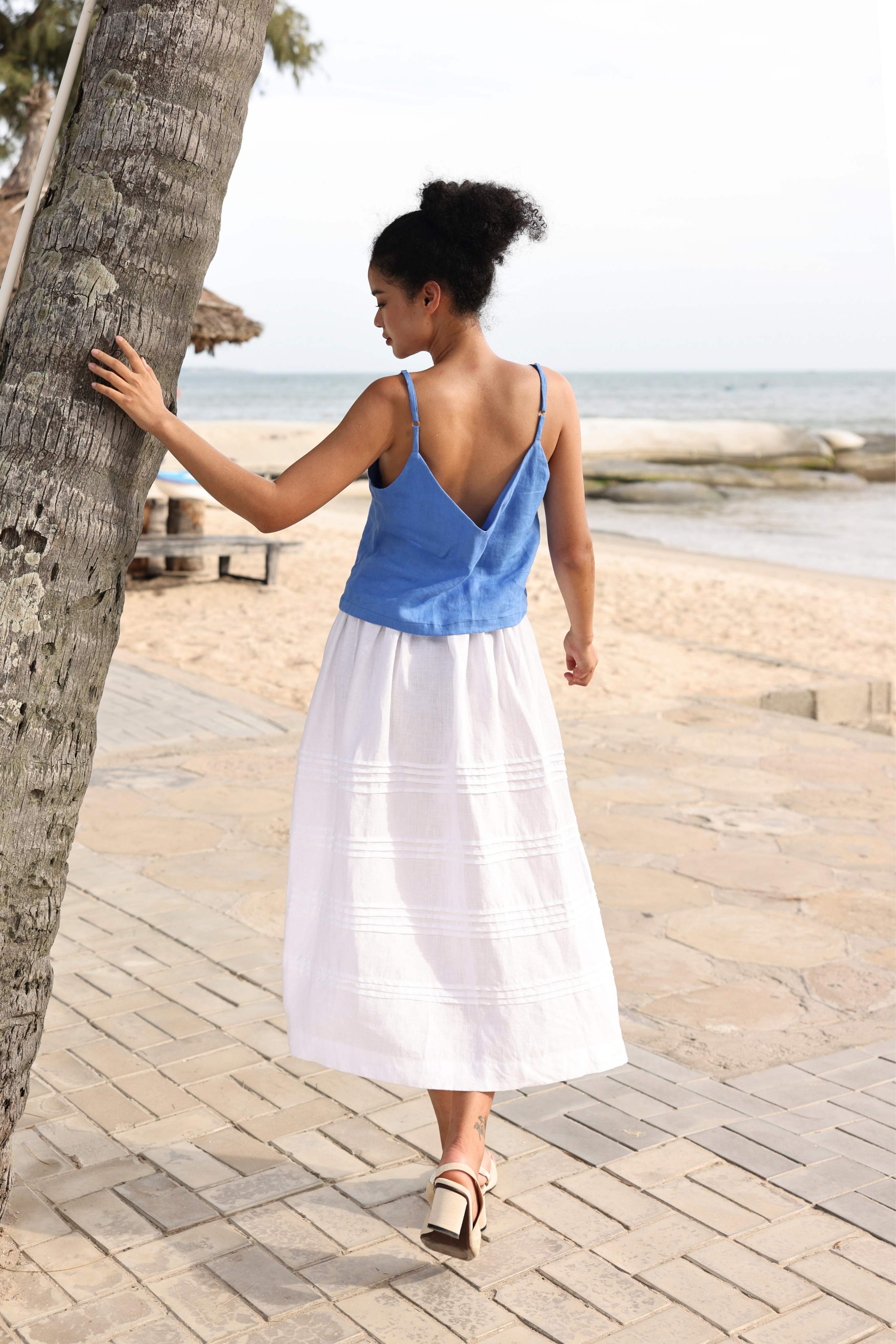 Woman in a blue top and white skirt walking along a wooden boardwalk by the beach.