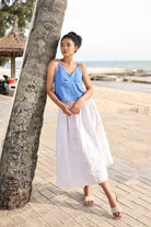 Woman in a blue top and white skirt standing by a tree on a beach.