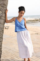 Woman in a blue top and white skirt standing by a palm tree on a beach.