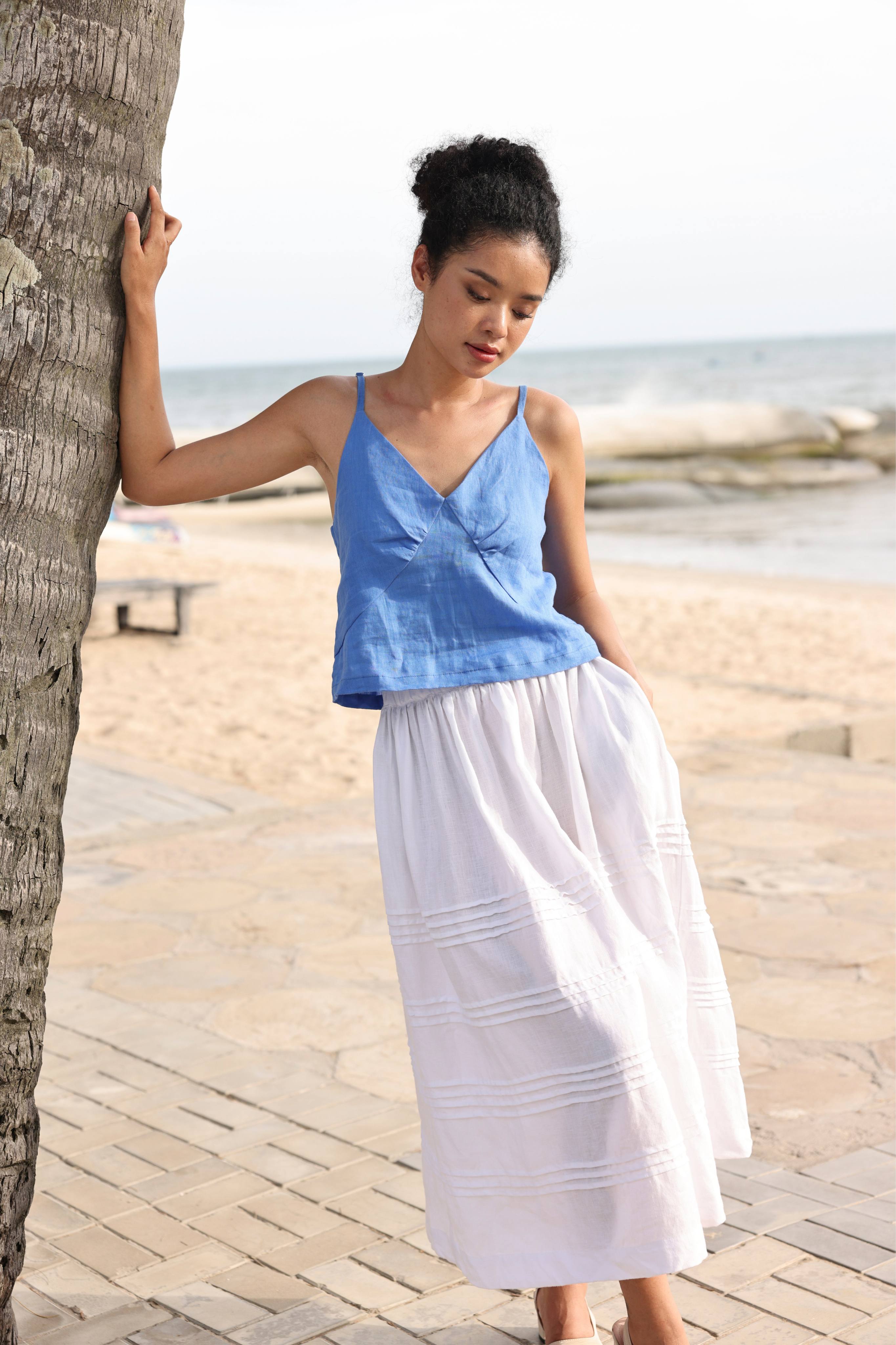 Woman in a blue top and white skirt standing by a palm tree on a beach.