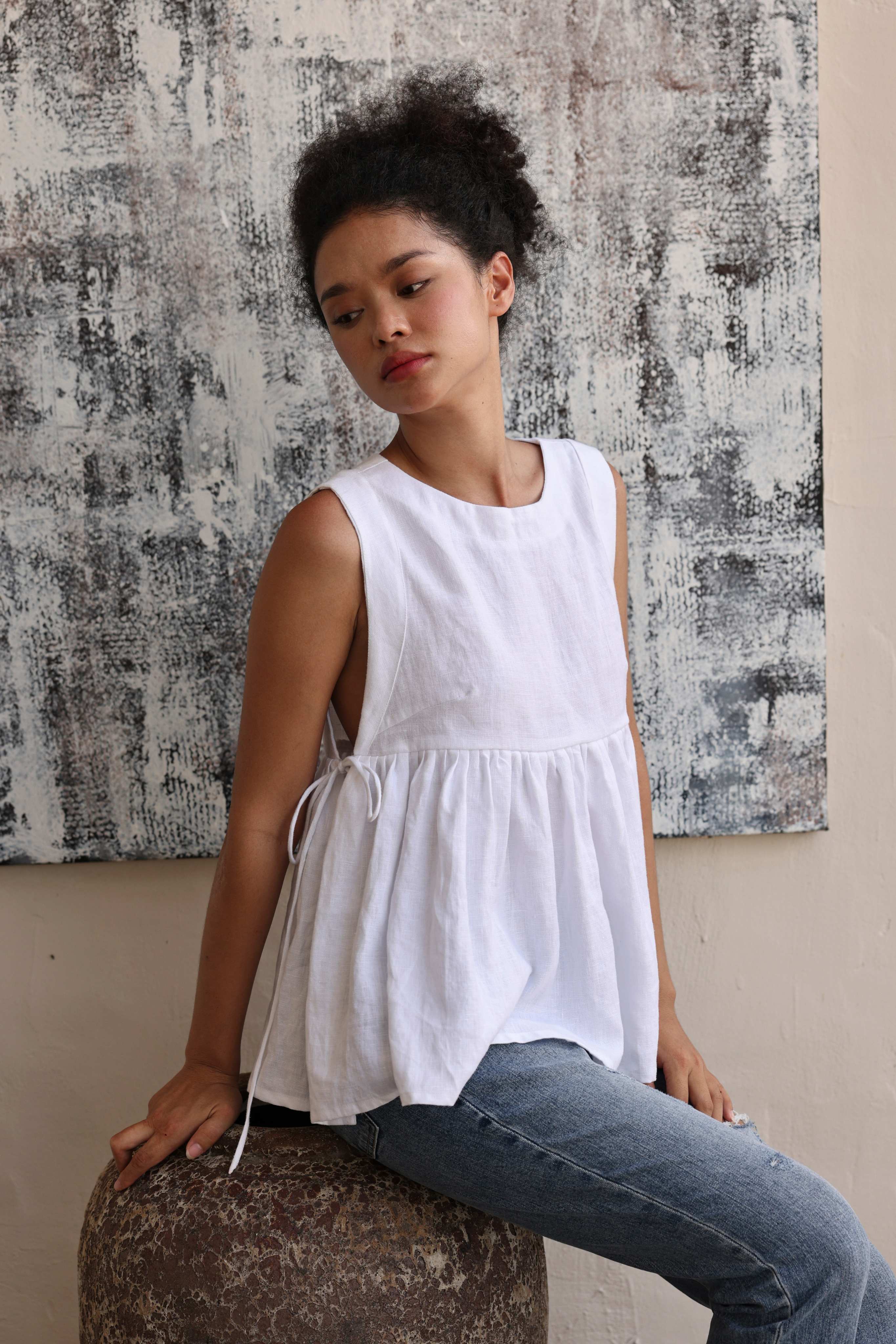 Woman wearing a white sleeveless top and jeans sitting on a textured surface with a patterned wall in the background.