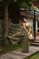 Woman in a green outfit standing on a wooden path with a hat in a garden setting.