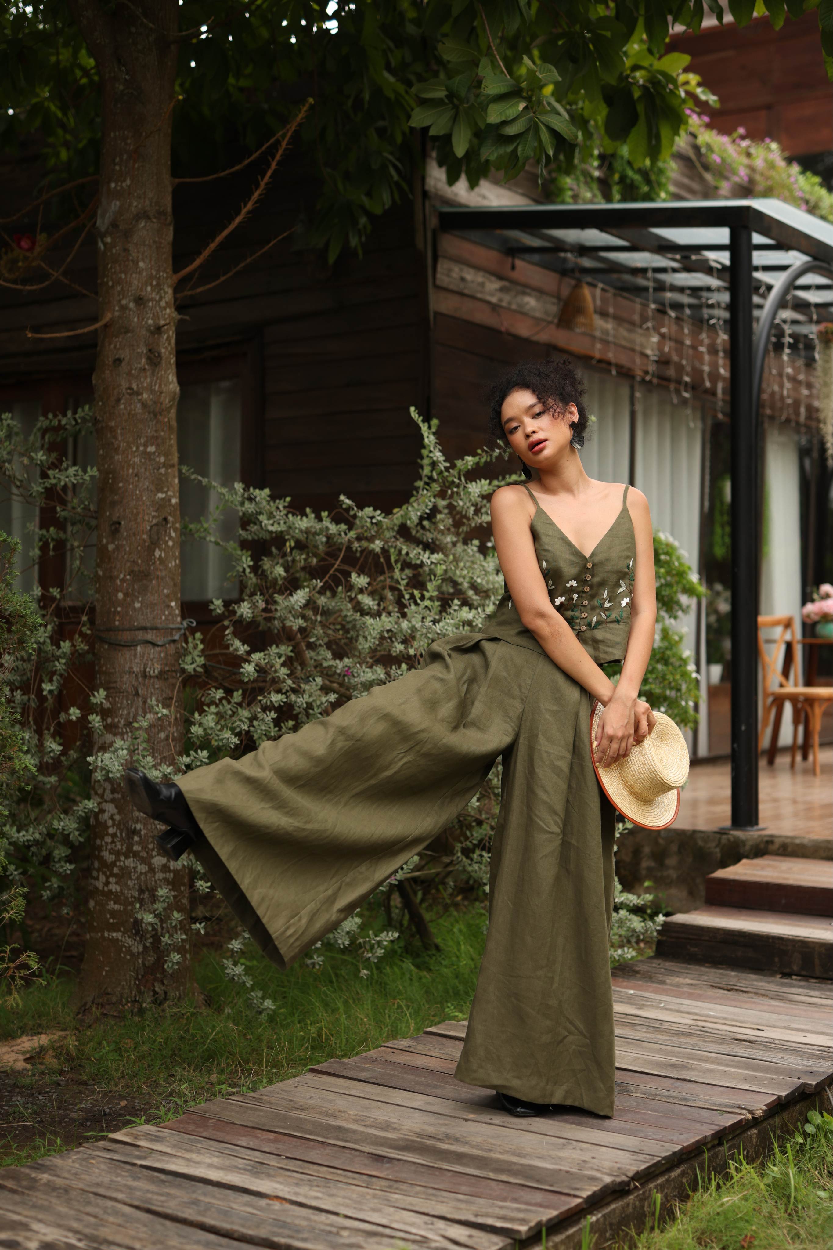 Woman in a green outfit standing on a wooden path with a hat in a garden setting.