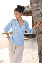 Woman in a light blue top and beige pants standing by a palm tree on a beach.