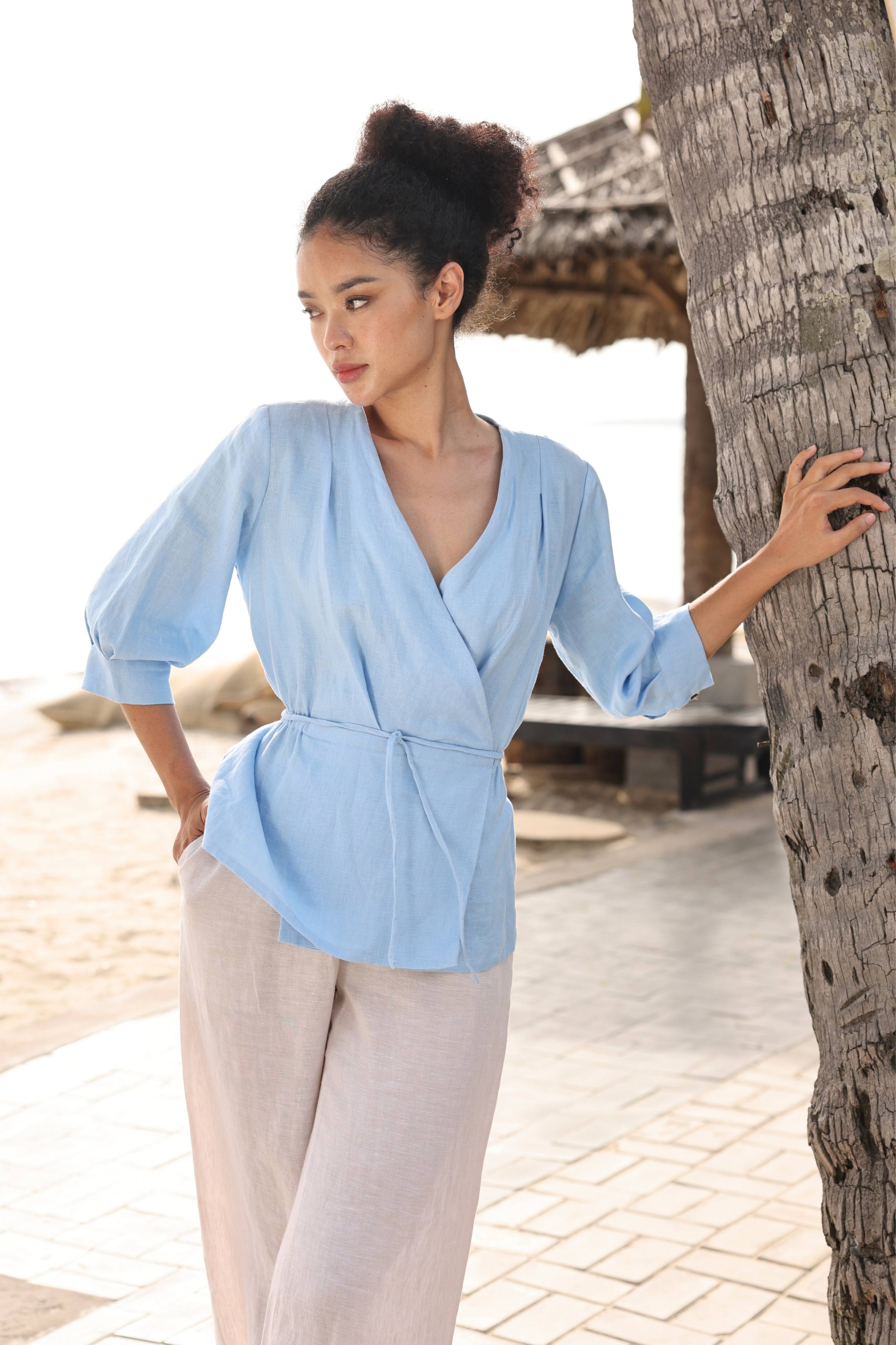 Woman in a light blue top and beige pants standing by a palm tree on a beach.