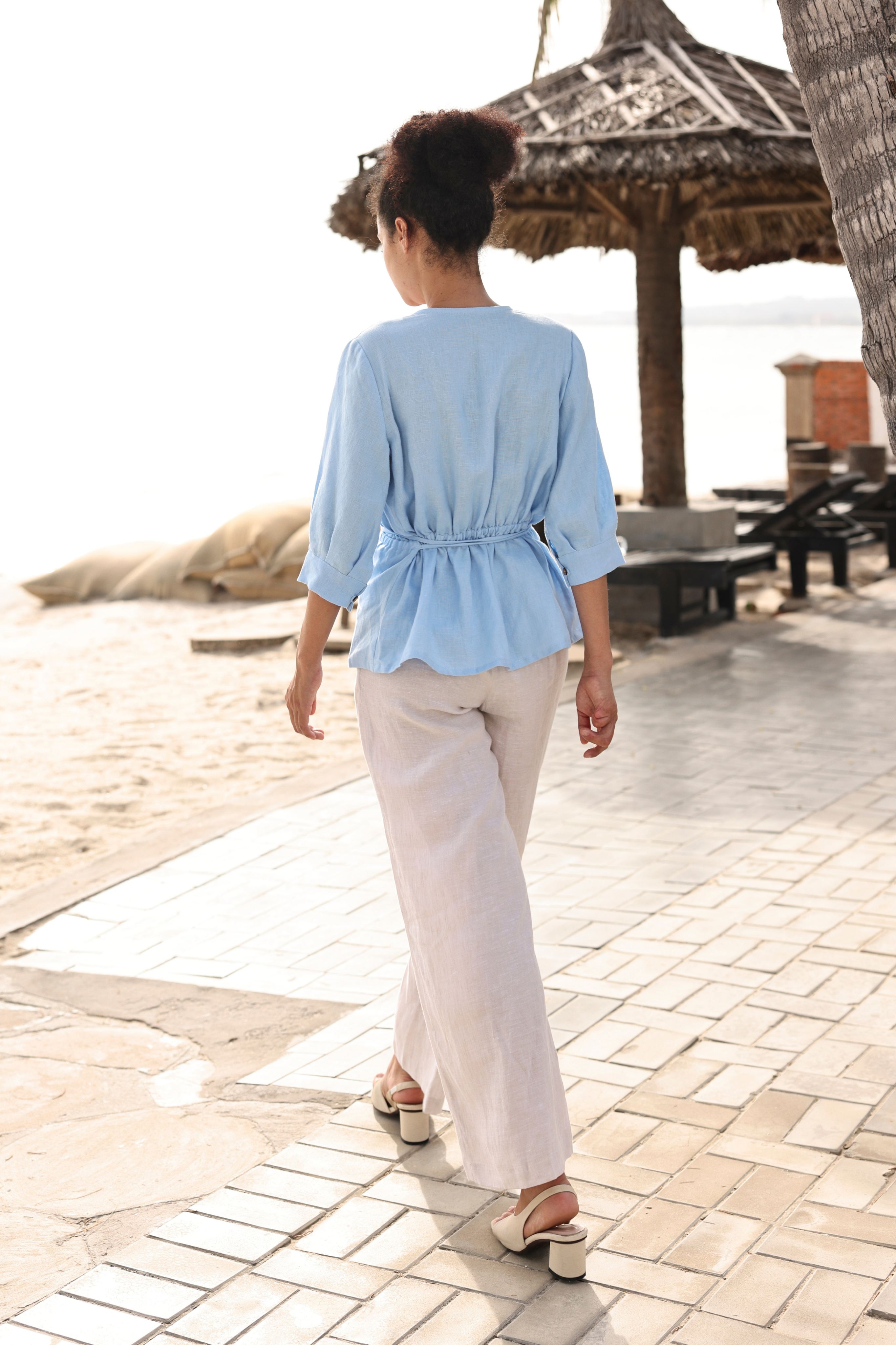 Person wearing a light blue top and white pants walking on a paved path near a beach with a thatched umbrella in the background.
