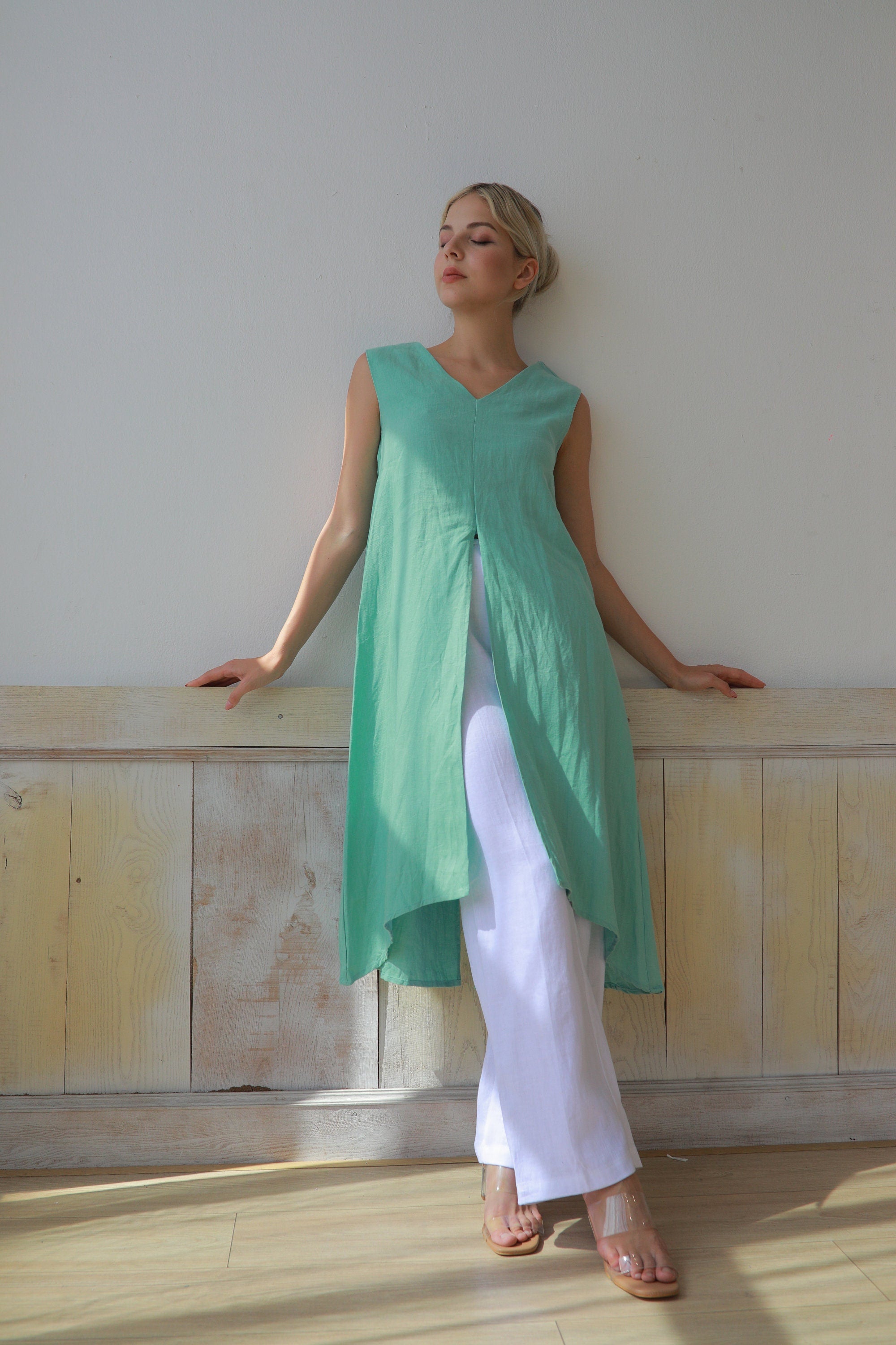 Woman wearing a green sleeveless tunic and white pants standing against a plain wall.