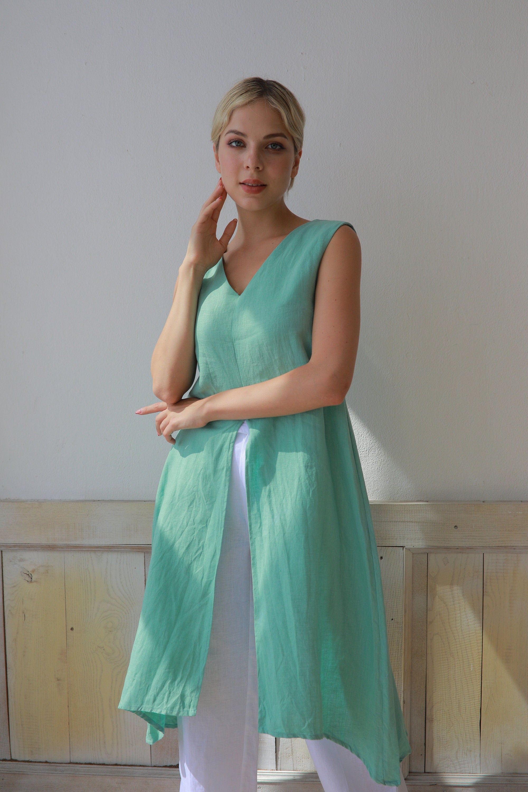 Woman wearing a light green sleeveless top and white pants against a plain wall.