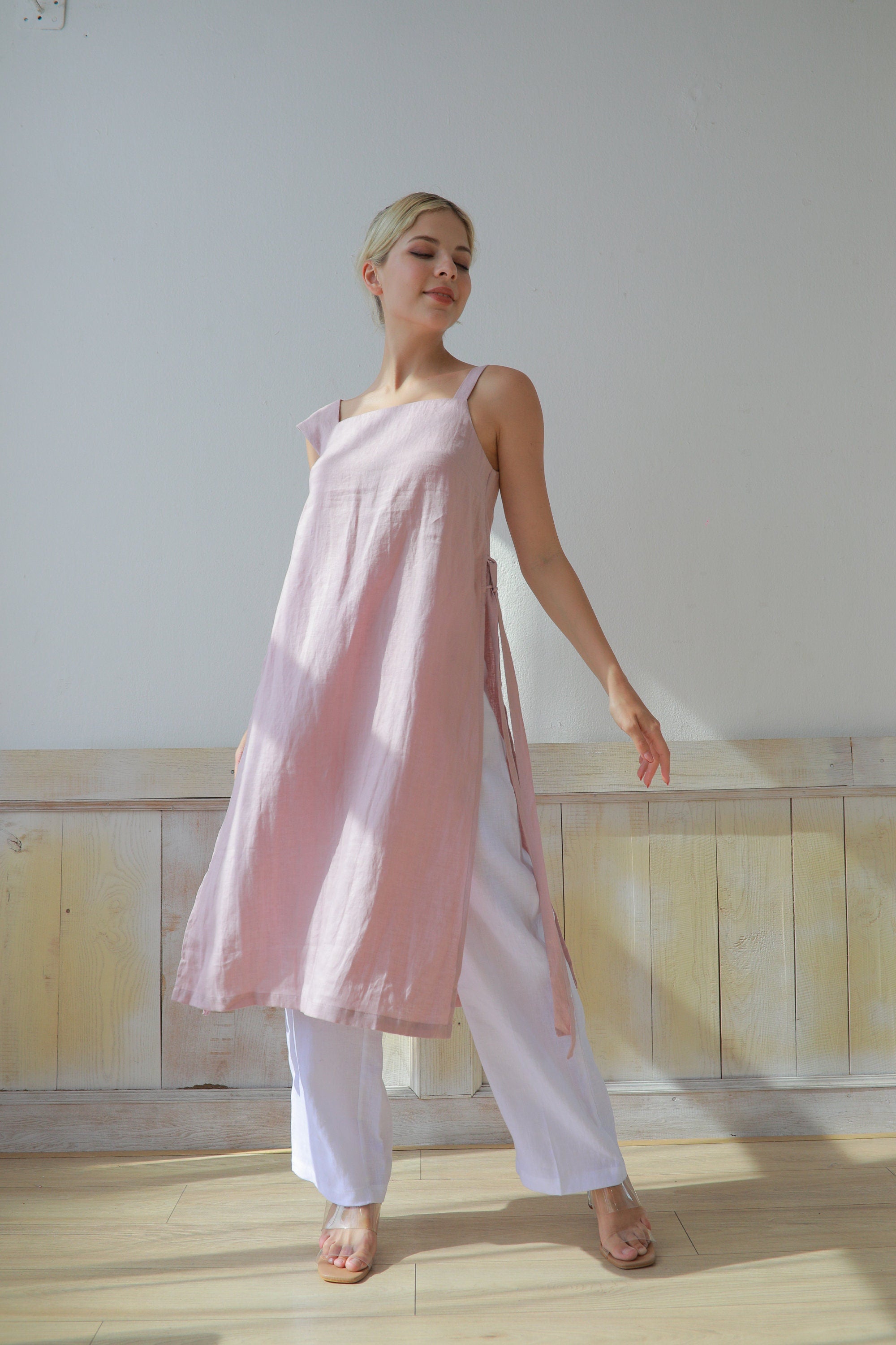 Woman wearing a pink dress and white pants standing in a room with wooden flooring and a light-colored wall.