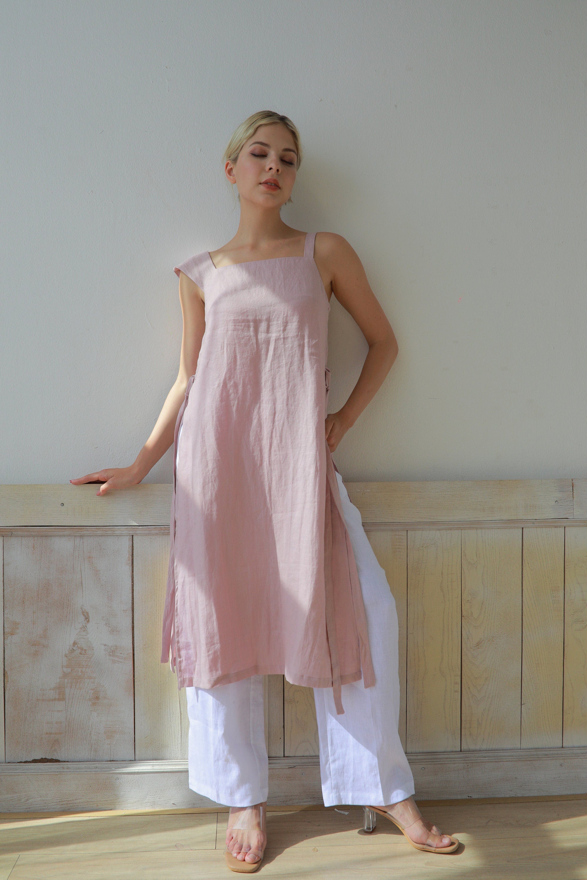 Woman wearing a pink dress and white pants standing against a plain wall.