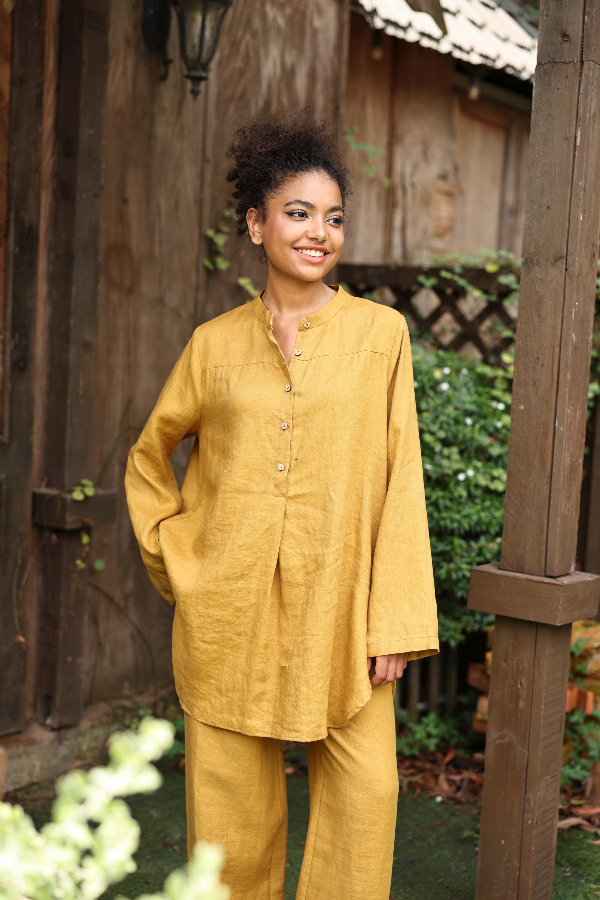 Woman wearing a mustard yellow outfit standing outdoors near wooden structures.