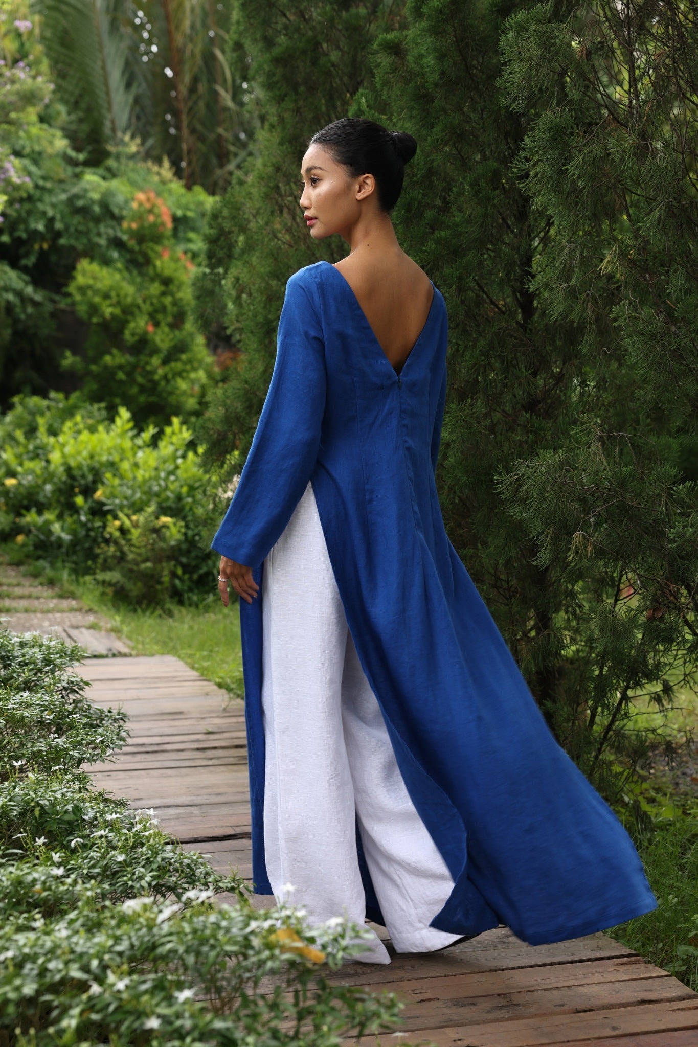 Woman in a blue tunic and white pants standing on a wooden path surrounded by greenery