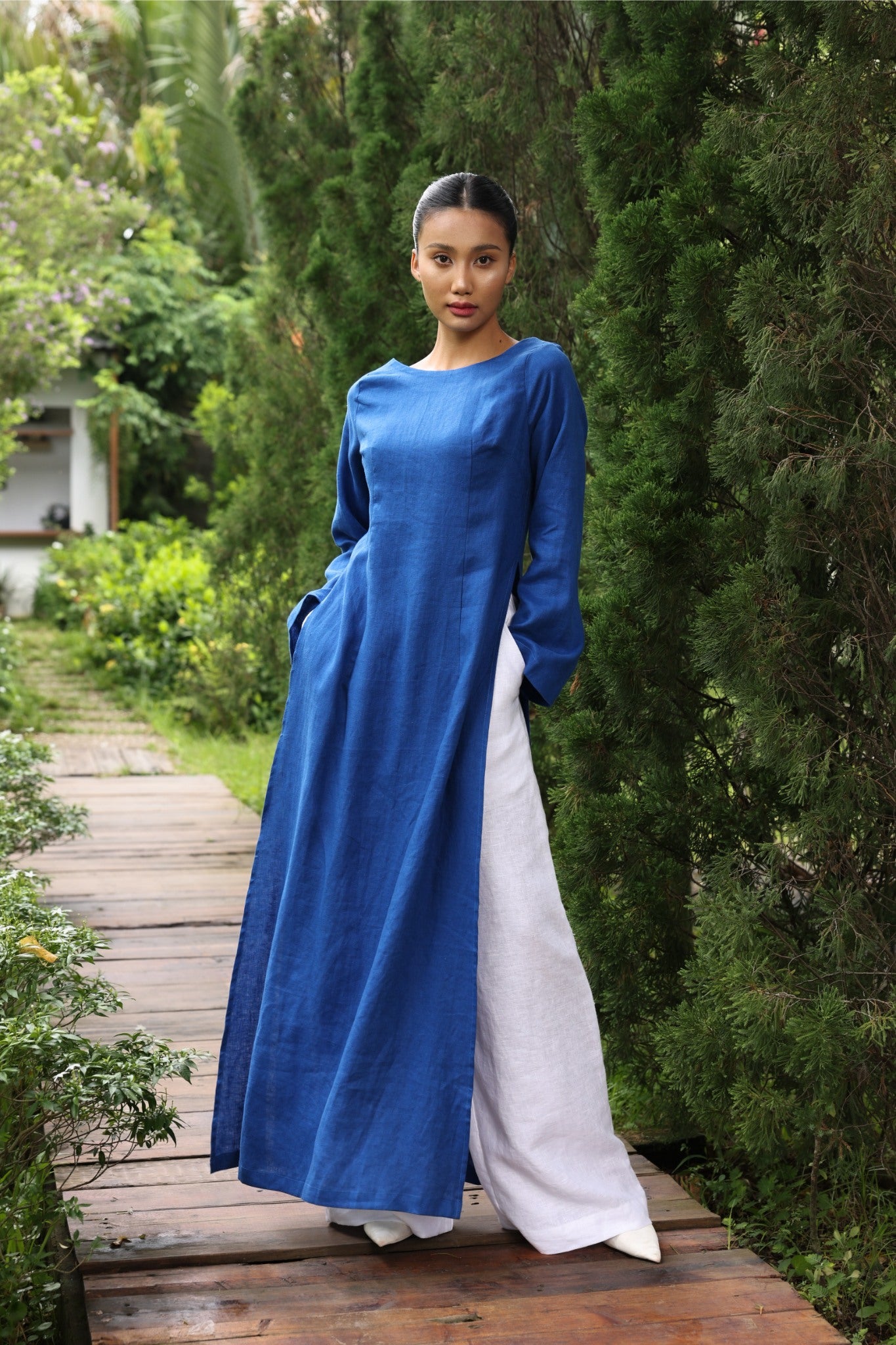 Woman wearing a blue tunic and white pants standing on a wooden path in a garden.
