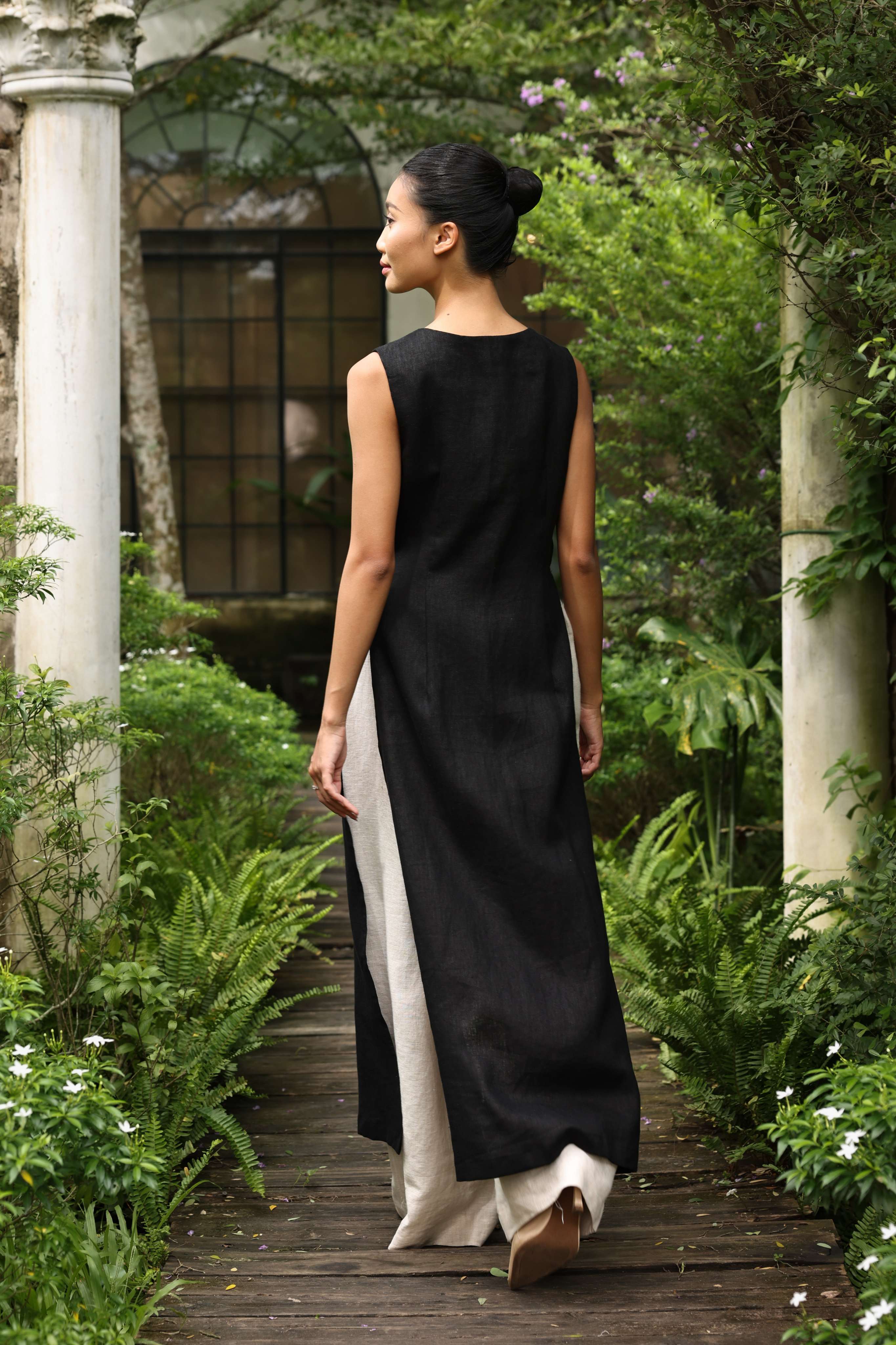 Woman in a black tunic and white pants walking through a garden with stone columns and greenery.