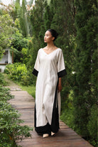 Woman in a long, light-colored dress with black trim standing on a wooden path in a garden.