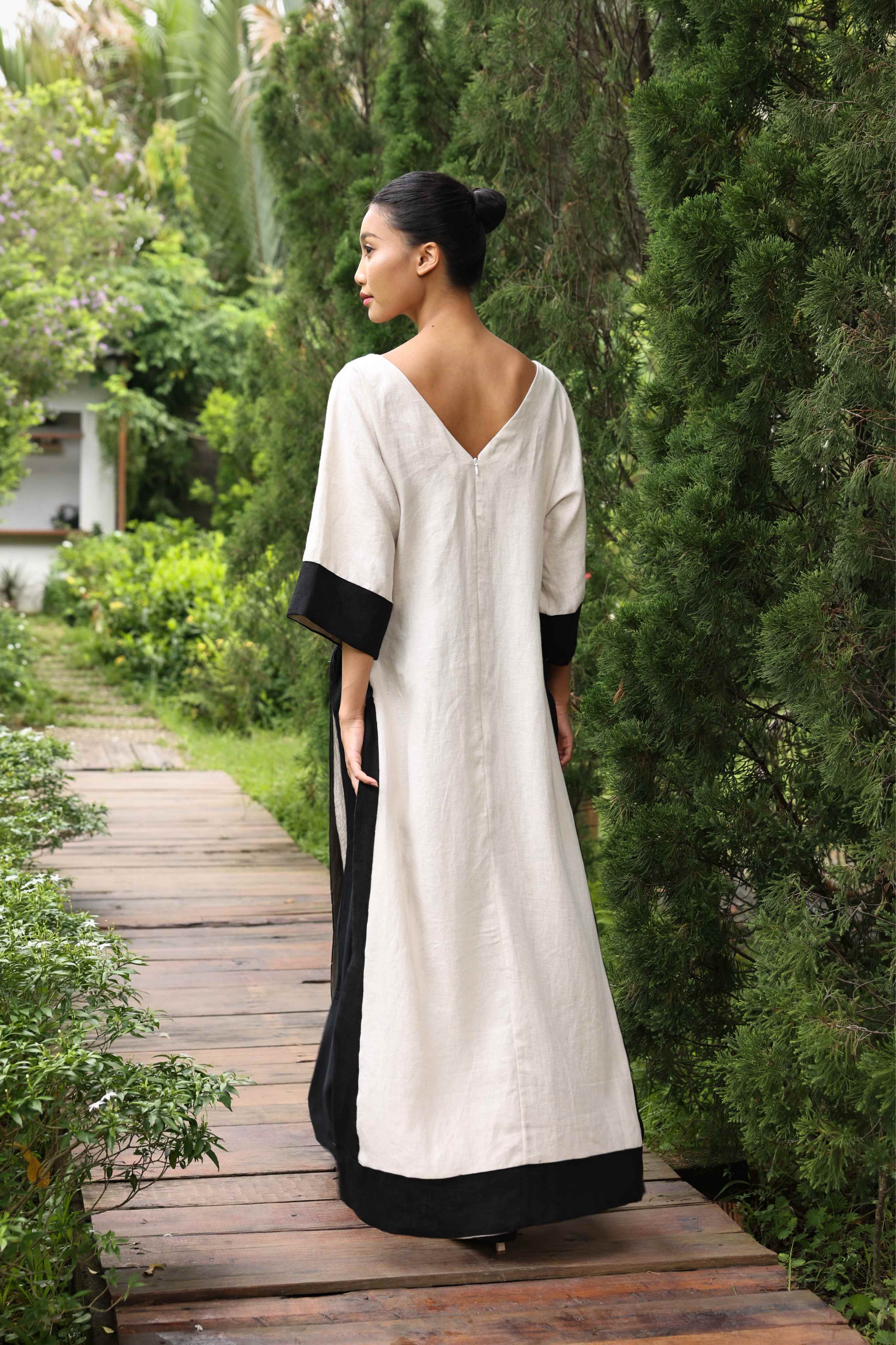Woman in a long, light-colored dress with black sleeves and trim walking on a wooden path in a garden.