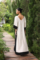 Woman in a long, light-colored dress with black sleeves and trim walking on a wooden path in a garden.