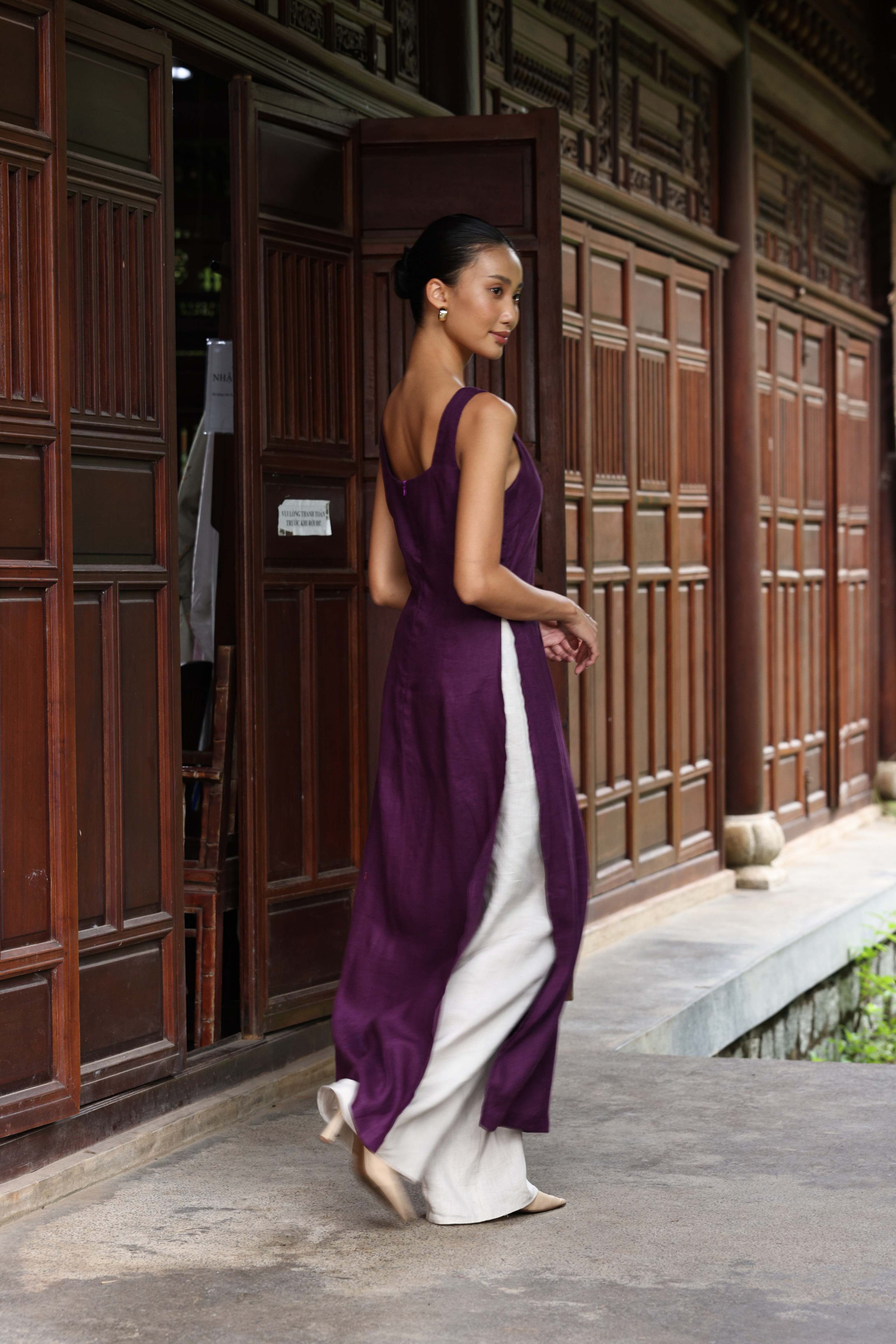 Woman in a purple tunic and white pants standing in front of a wooden building.