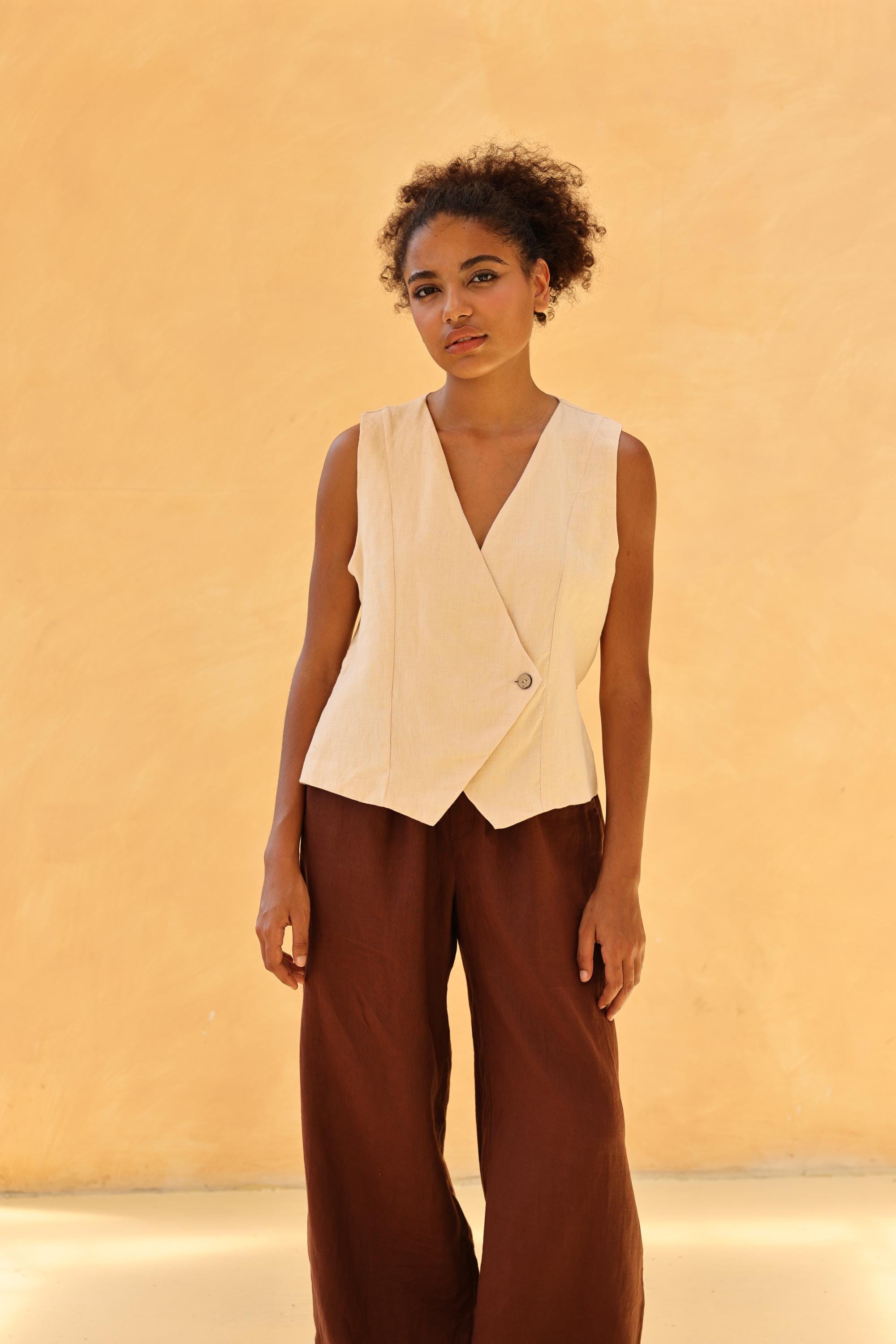 Woman wearing a sleeveless blazer and brown pants against a beige background